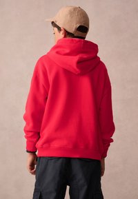 Červená mikina s kapucí a uvolněným střihem, s žebrovanými manžetami a lemem. Na hlavě je béžová čepice, která doplňuje volnočasový outfit.