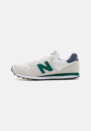 Witte en beige sneaker met groen "N"-logo, navy blauwe hielband, witte veters en zwarte textuurzool, zijaanzicht.