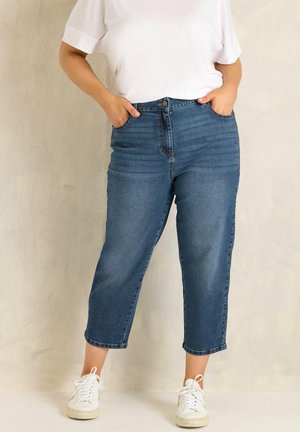 Persona che indossa jeans blu corti con vita alta, t-shirt bianca e sneakers bianche, in piedi con le mani nelle tasche davanti a uno sfondo semplice.