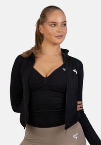 Zwarte sportjack met ritssluiting, slim fit ontwerp, zachte stof en lange mouwen, gelaagd over een zwart topje. Beige high-waisted leggings.