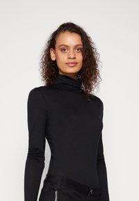Black long-sleeve turtleneck top made of smooth, stretchy fabric, featuring a fitted design and a high collar, paired with dark slim-fit pants.
