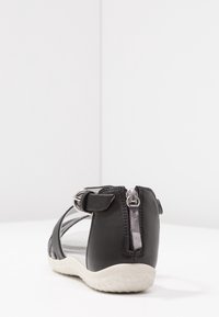 Musta nahkasandaali, jossa on nilkkaremmu, takana vetoketju ja teksturoitu valkoinen kumipohja, nähty vasemman takakulman näkökulmasta valkoisella pinnalla.