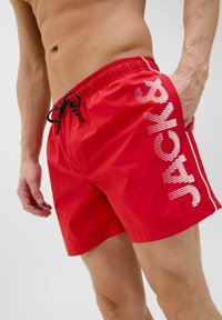 Bañador rojo hecho de tejido ligero, con un cordón de ajuste negro, rayas laterales blancas y un texto en negrita en la parte delantera.