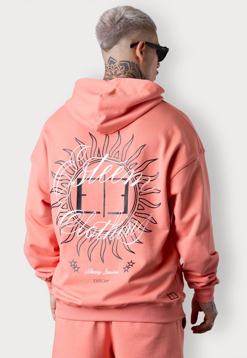 Un sweat à capuche rose corail avec une coupe décontractée, mettant en avant un graphique de soleil et le texte "Blazing Sunshine" en noir et blanc. Poignets et ourlet côtelés.