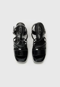 Fekete lakkbőr magassarkú szandál, többpántos dizájnnal, nyitott orral, négyzet alakú kialakítással és állítható boka csattal. Szív logó a talpbetéten.