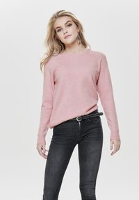 Helles rosa Sweatshirt mit Rundhalsausschnitt und ribbelten Bündchen, kombiniert mit schwarzen Skinny-Jeans und einem Nieten-Gürtel. Glatte Textur, lässiges Design.
