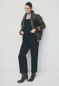 Schwarze Leder-Bomberjacke über einem grauen Pullover, kombiniert mit schwarzen Overalls und burgunderroten Stiefeletten mit Absatz. Glatte Texturen, schlankes Design.