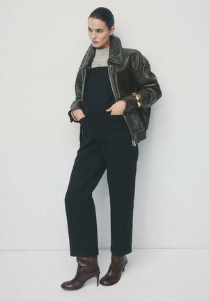 Schwarze Leder-Bomberjacke über einem grauen Pullover, kombiniert mit schwarzen Overalls und burgunderroten Stiefeletten mit Absatz. Glatte Texturen, schlankes Design.