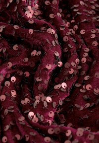 Burgunderfarbener Stoff mit reflektierenden Pailletten in runden Formen, die eine strukturierte Oberfläche mit wechselnden glänzenden und matten Flächen schaffen.