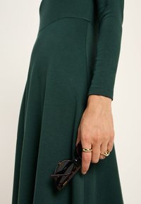 Hand, die eine Schildpatt-Sonnenbrille hält, trägt ein dunkelgrünes langärmliges Kleid und zwei goldene Ringe an den Fingern.