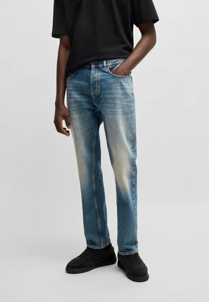 Personne portant un jean bleu délavé, des baskets noires et un t-shirt noir avec une main dans la poche sur un fond uni.