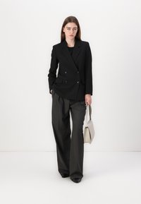 Černý dvouřadý blazer s knoflíky v kombinaci se šedými širokými kalhotami s jemným proužkem. V jedné ruce drží světle béžovou kabelku.