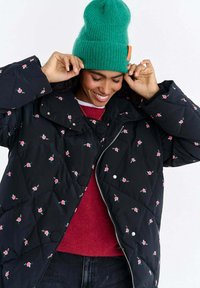 Czarna pikowana kurtka w kwiatowy wzór, z zapięciem na zatrzaski i zamek błyskawiczny. Noszona na czerwonym topie i zestawiona z zieloną, dzianą czapką beanie.