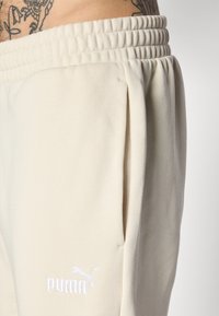 Pantalons de survêtement beige clair avec une ceinture côtelée, des poches latérales et un logo PUMA brodé en blanc sur la cuisse gauche. Tissu doux et texturé.