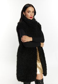 Zwart harig faux fur zonder mouwen vest, hoge kraag, gecombineerd met een strak zwart langslijvig top en een gouden metallic minirok. Zwart knielaarzen.