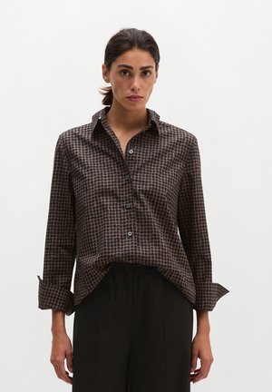 Vrouw met donker haar draagt een bruin patroon overhemd met knopen en zwarte broek, staand tegen een effen lichte achtergrond.