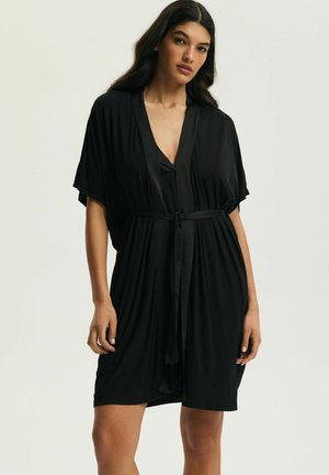 Žena s dlhými tmavými vlasmi, oblečená v čiernom župane po kolená zviazanom v páse, stojaca pred jednoduchým svetlým pozadím.