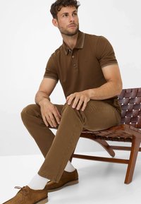 Polo shirt marrone con colletto e polsini a contrasto. Pantaloni marroni slim-fit, scarpe in camoscio marroni, seduto su una sedia di legno con dettagli intrecciati.