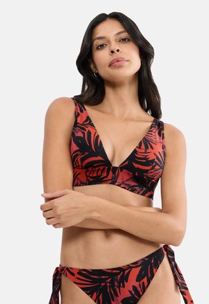 Kobieta w czerwono-czarnym bikini z tropikalnym wzorem, zawiązanymi bokami dołu, z założonymi ramionami i długimi ciemnymi włosami na jednolitym tle.