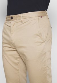 Chinos in cotone beige con una texture liscia, taglio dritto e tasche frontali. Presentano un bottone marrone e un piccolo accento del marchio multicolore.