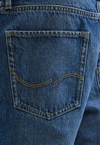 Jean en denim avec une poche arrière ornée de coutures jaunes et d'un motif de couture décoratif courbé sur un tissu bleu.