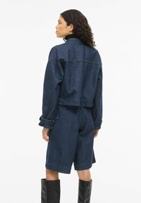 Tumši zila džinsa jaka ar īsu dizainu, kopā ar atbilstošiem šortiem. Raksturīgas platās piedurknes un uzlocītie aproces gali. Melni ceļgalus noklājoši zābaki papildina izskatu.