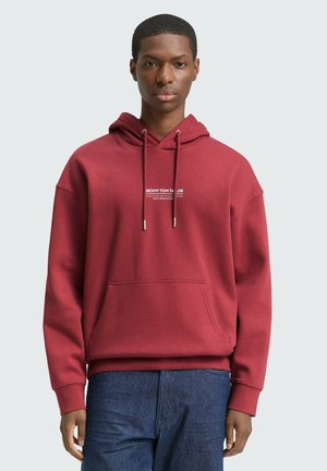 Maroon Kapuzenpullover aus weichem Stoff mit einer Kängurutasche und Kordelzug-Kapuze. Textbranding ist auf der Vorderseite aufgedruckt. Jeans aus Denim sind sichtbar.