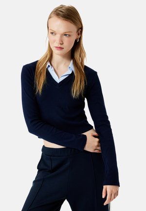 Jonge vrouw met lang blond haar die een cropped navy V-hals trui draagt over een blauw gestreept shirt en marineblauwe broek, kijkt zijwaarts.