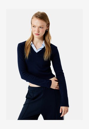 Jeune femme aux longs cheveux blonds portant un pull court bleu marine à col en V par-dessus une chemise bleue à rayures et un pantalon bleu marine, regardant sur le côté.