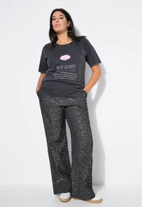 Grijze t-shirt met grafische print en tekst, gecombineerd met zwarte broek met wijde pijpen en zilveren stippenpatroon. Witte sneakers maken de look af.