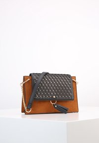 Hnědá semišová kabelka s černou geometrickou klopou, zlatými kovovými prvky a odnímatelným řetízkem. Obsahuje přívěsek s třásněmi.