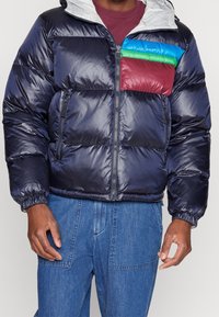 Doudoune matelassée bleu marine avec une finition brillante. Dispose d'une section poitrine bicolore en bordeaux, vert et bleu, avec des poignets élastiques et une capuche.