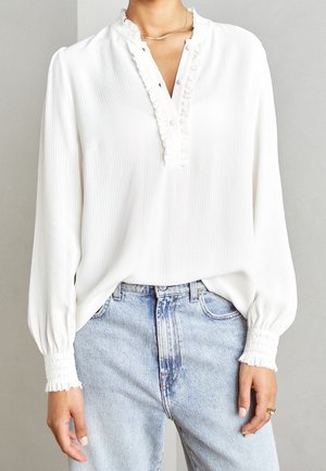 Mujer que lleva una blusa blanca de manga larga con cuello y puños con volantes, combinada con vaqueros de talle alto de color azul claro y un collar fino de plata.