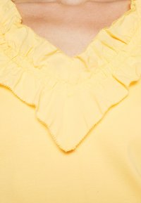 Gele blouse met een gerimpelde halslijn met golvende randen. Zachte, lichte stof met een fijne textuur. Geen extra patronen of accenten.