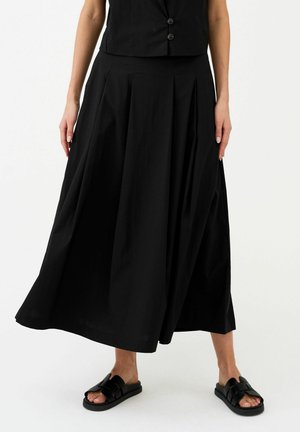 Vrouw draagt een zwarte mouwloze top, zwarte wijde cropped broek en zwarte slippers, staand op een witte achtergrond.