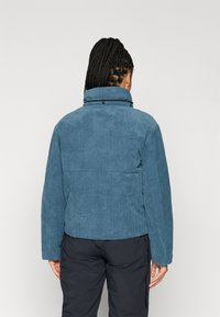 Modrá manšestrová bunda s vysokým límcem a uvolněným střihem, s vertikálními proužky a patentkami u krku. Viditelné černé kalhoty.