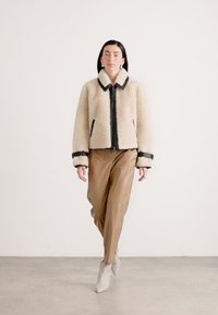 Beige schapenvachtjas met zwarte afwerking, volledige rits en zijzakken, gecombineerd met tan leren broeken en beige enkellaarzen.
