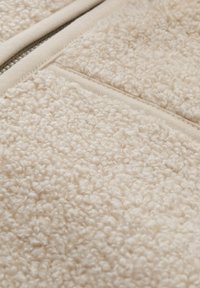 Tissu en polaire beige avec une surface douce et texturée, featuring une fermeture éclair lisse couleur fauve et un ourlet côtelé contrastant le long des bords.