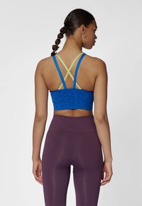 Blå sport-bh med gröna korslagda band, med ett texturerat mönster. Matchas med högmidjade lila tights.