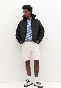 Mann trägt schwarze Jacke, blau-weiß gestreiftes Hemd, weiße Shorts, weiße Turnschuhe, mit Tätowierungen an den Beinen, steht vor weißer Wand.