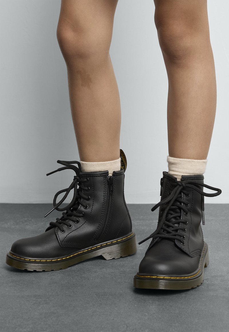 Jambes portant des bottines en cuir noir à lacets avec fermeture éclair latérale et chaussettes beige sur un sol gris contre un mur gris.