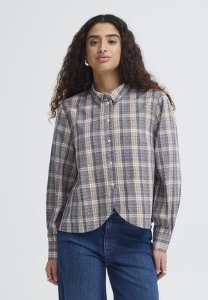 Jeune femme aux cheveux longs et bouclés portant une chemise à carreaux grise et bleue boutonnée et un jean bleu, sur un fond uni.