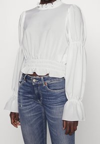 Blouse blanche à manches longues avec un col et des poignets volantés, taille cintrée, associée à un jean en denim bleu foncé. Tissu lisse et coupe décontractée.