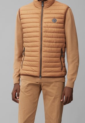 Homme portant une veste matelassée beige zippée sur un pull beige à manches longues et un pantalon beige, debout devant un fond gris.
