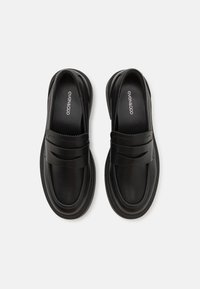 Fekete bőr, slip-on stílusú cipők lekerekített orral, díszítő pánttal és fodros részletekkel a felső részen.