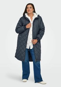 Prošívaný námořnický dlouhý kabát s kapucí, který má bílou sherpa podšívku, přední zip a nastavitelné stahovací šňůrky. Na sobě jsou vykrojené džíny a bílé boty.