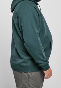 Homme portant un sweat à capuche vert et un pantalon gris, debout de profil sur un fond uni.