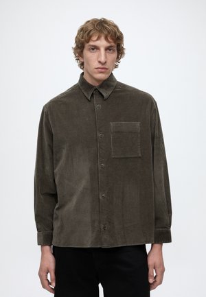 Homme aux cheveux blonds bouclés portant une chemise en velours côtelé vert olive foncé à boutons avec une poche poitrine et un pantalon noir.