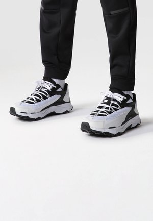 Person trägt schwarz-weiße Sportschuhe mit weißen Schnürsenkeln, weiße Socken und schwarze Hosen und steht auf einer weißen Oberfläche.