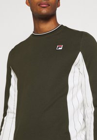 Camiseta de manga larga verde oliva con paneles laterales de rayas blancas y cuello de canalé, que cuenta con un pequeño parche de logo en el pecho.